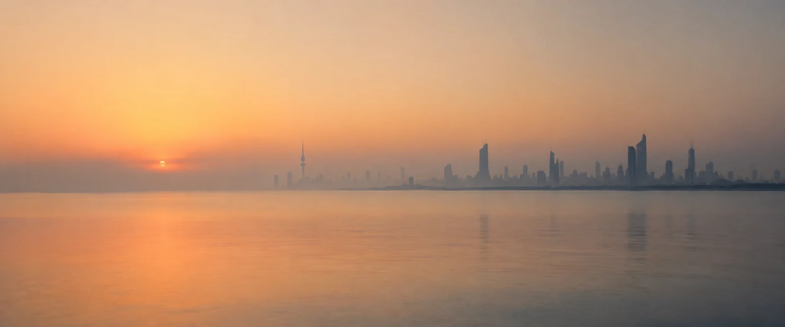 Kuwait City skyline silhouetted against a hazy orange sunrise over calm water