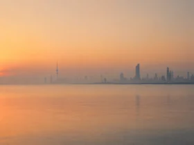 Kuwait City skyline silhouetted against a hazy orange sunrise over calm water