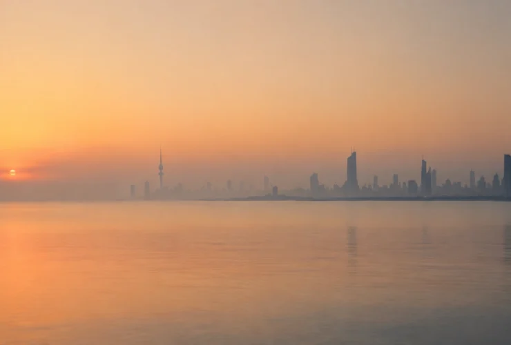 Kuwait City skyline silhouetted against a hazy orange sunrise over calm water
